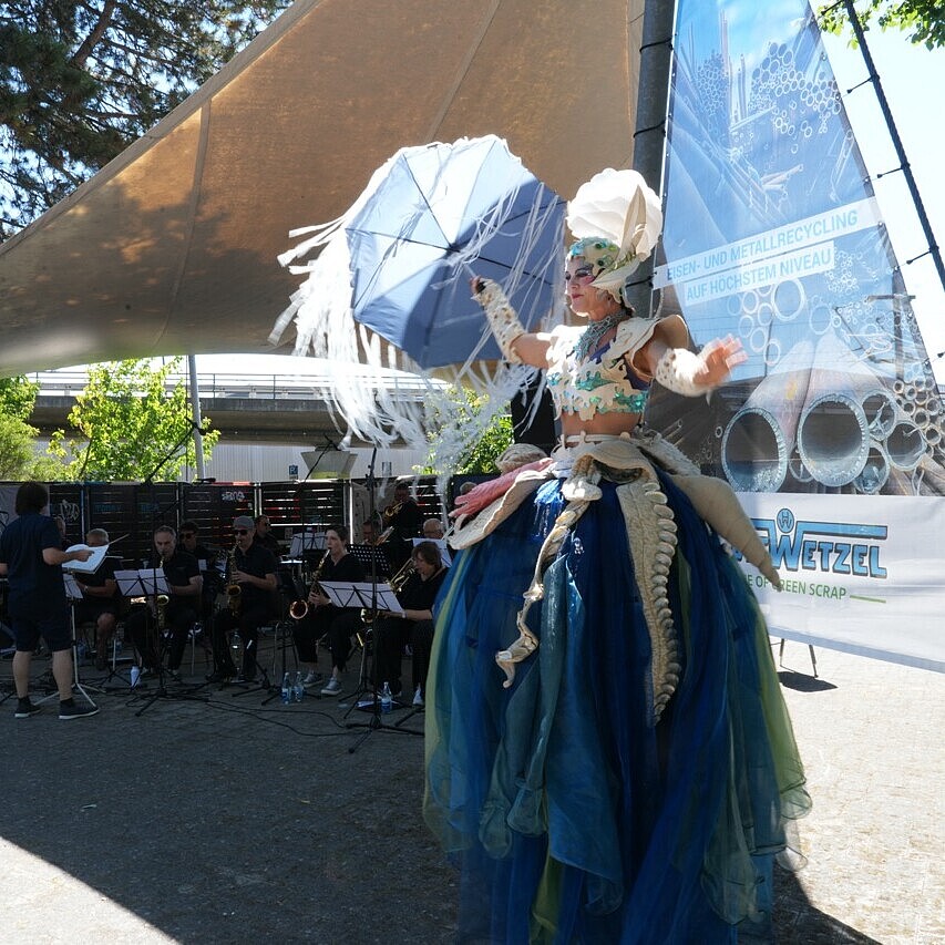 Stelzenläuferin als Walking Act beim HafenKulturFest 2025 am Karlsruher Rheinhafen, mit Publikum entlang der Kaimauer und Industriekran im Hintergrund.
