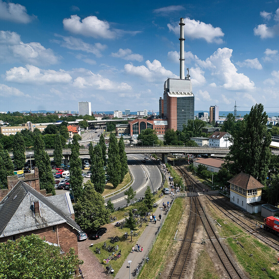 Blick auf Bahngleise im Rheinhafen Karlsruhe mit Industriegebäuden, Straßenführung und städtischer Umgebung bei Tageslicht.