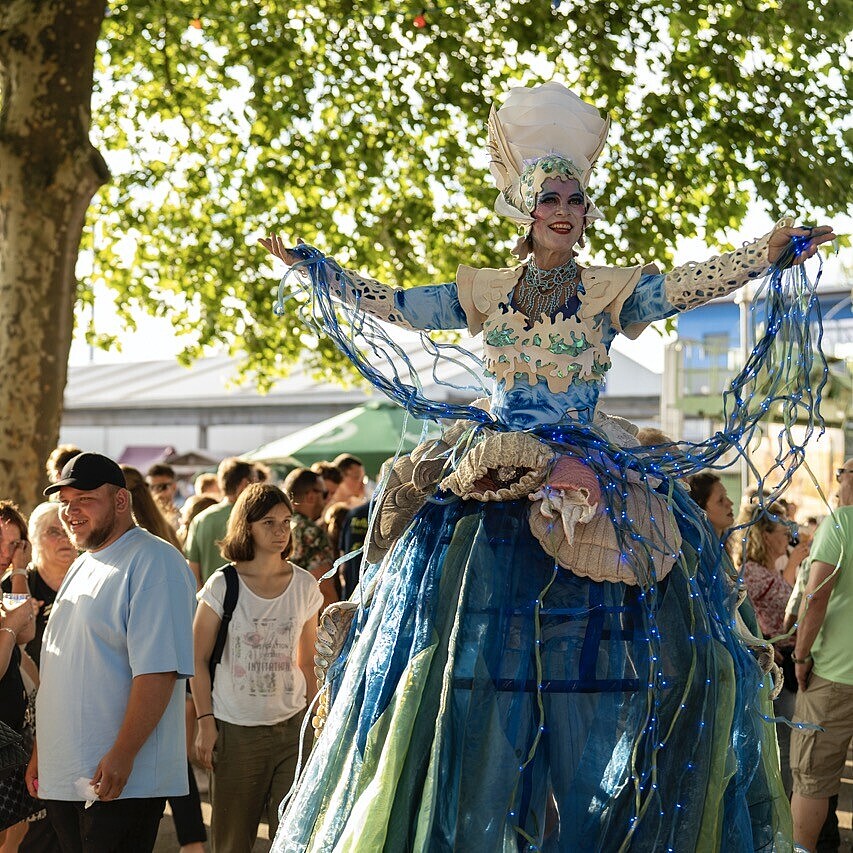 Stelzenläuferin als Walking Act beim HafenKulturFest 2025 am Karlsruher Rheinhafen, mit Publikum entlang der Kaimauer und Industriekran im Hintergrund.