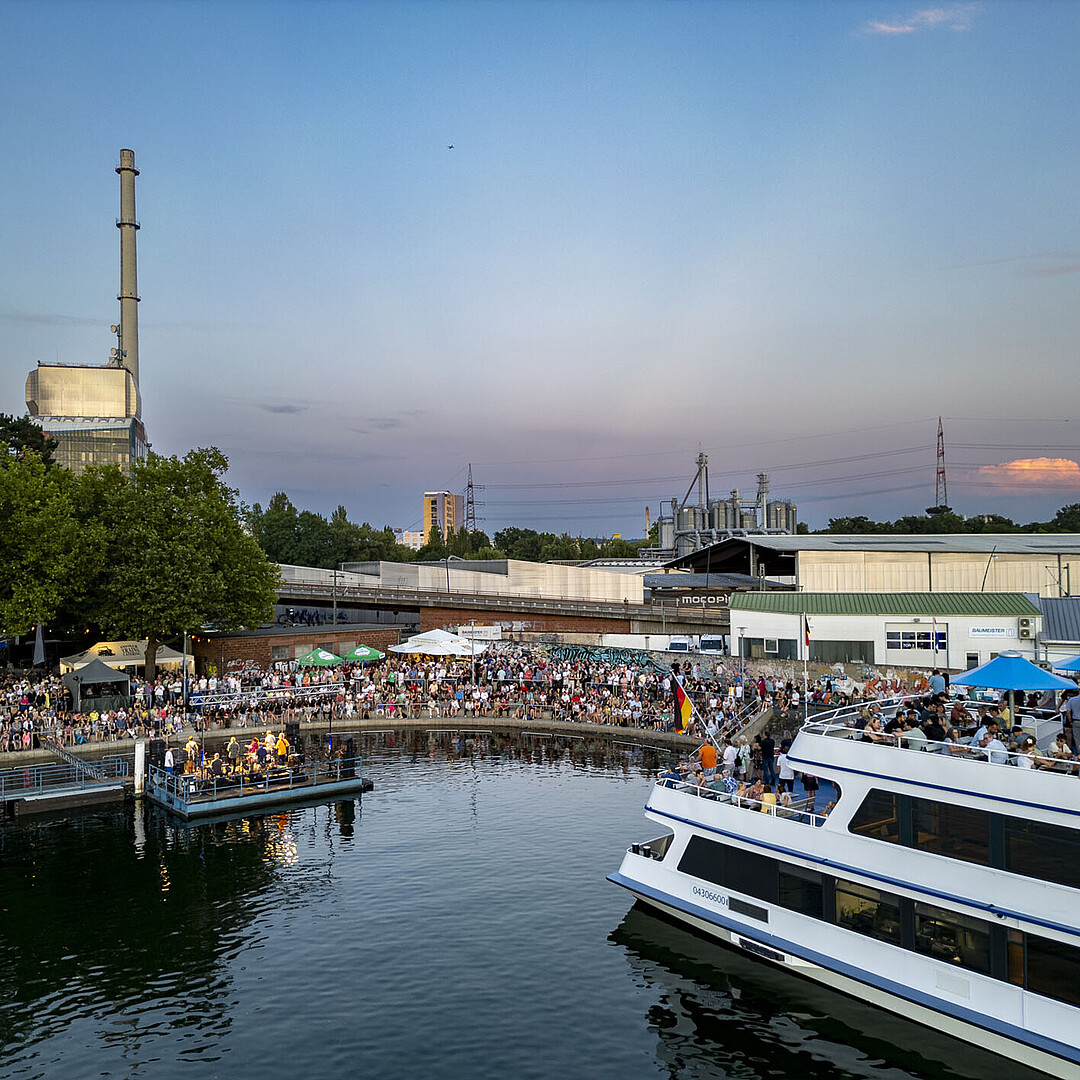 Fahrgastschiff MS Karlsruhe am Anleger im Rheinhafen Karlsruhe bei Abendstimmung, mit Gästen an Bord und beleuchtetem Hafenumfeld.